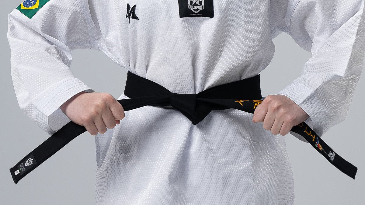 Close-up das m&atilde;os de uma atleta brasileira amarrando a faixa preta sobre o dobok Sulsport com bandeira do Brasil no ombro