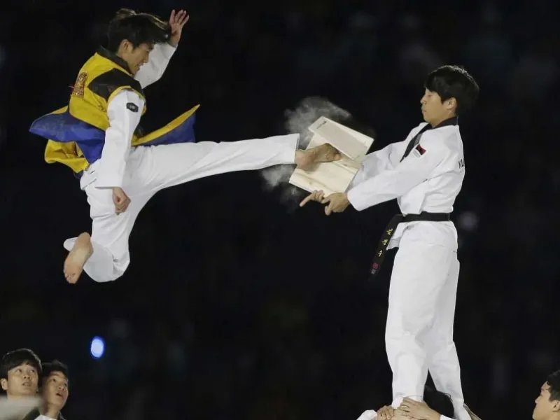 Atleta de Taekwondo executando chute voador para quebrar t&aacute;bua de madeira em demonstra&ccedil;&atilde;o, com part&iacute;culas de madeira voando ao impacto